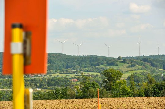 Schilderpfahl an einem Feld mit Waldrand und Windrädern im Hintergrund